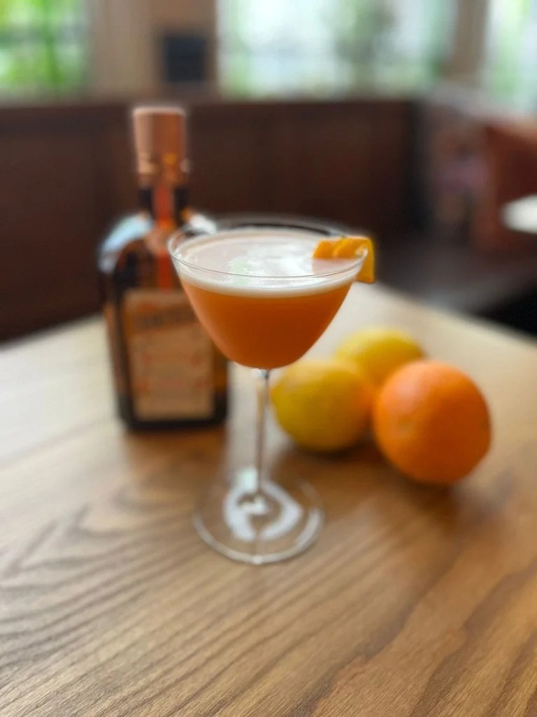 An orange cocktail in a coupe glass with citrus and a bottle behind it